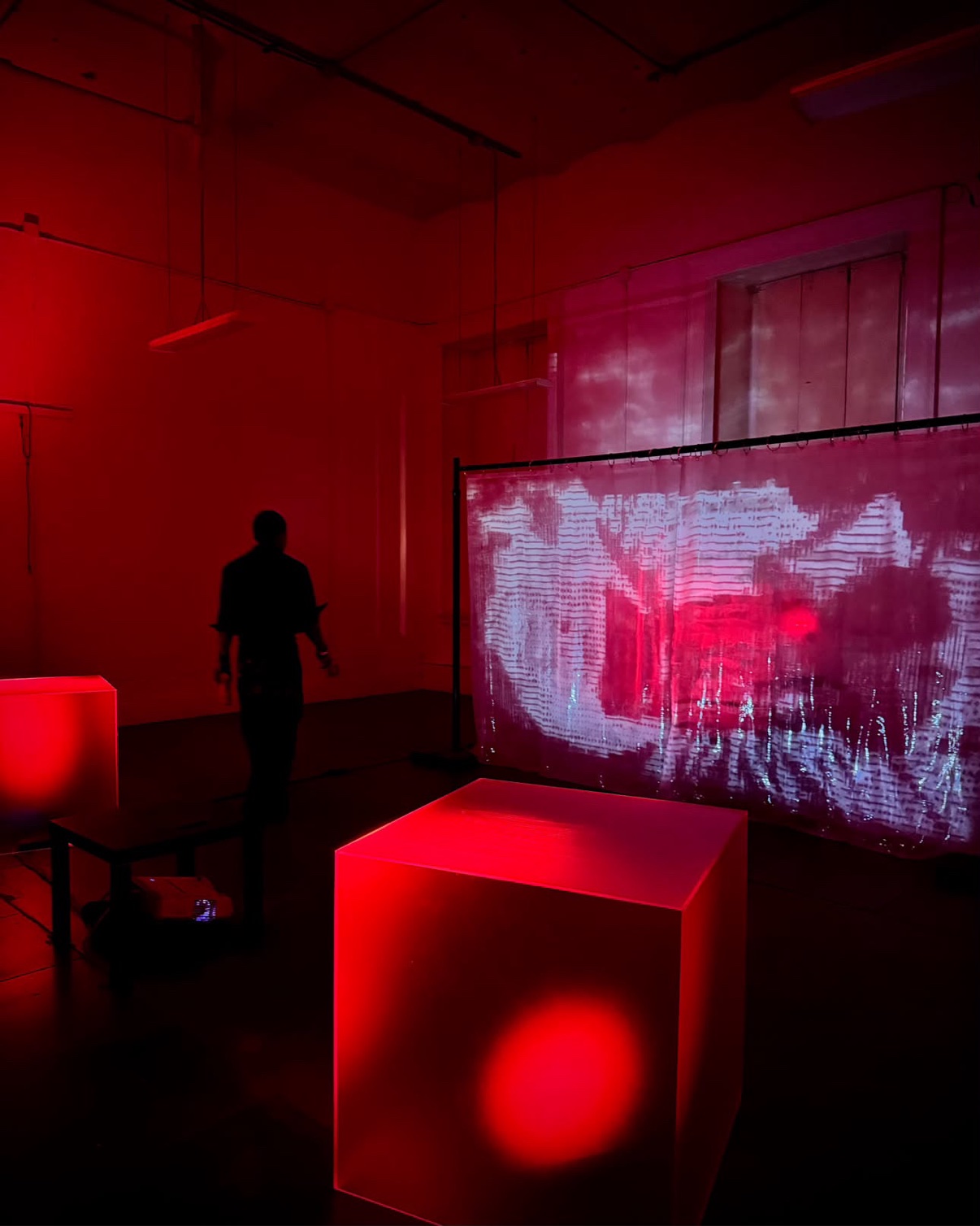 The art installation room washed in red light — glowing red cubes and a projection playing across a translucent curtain, a silhouetted figure walking through
