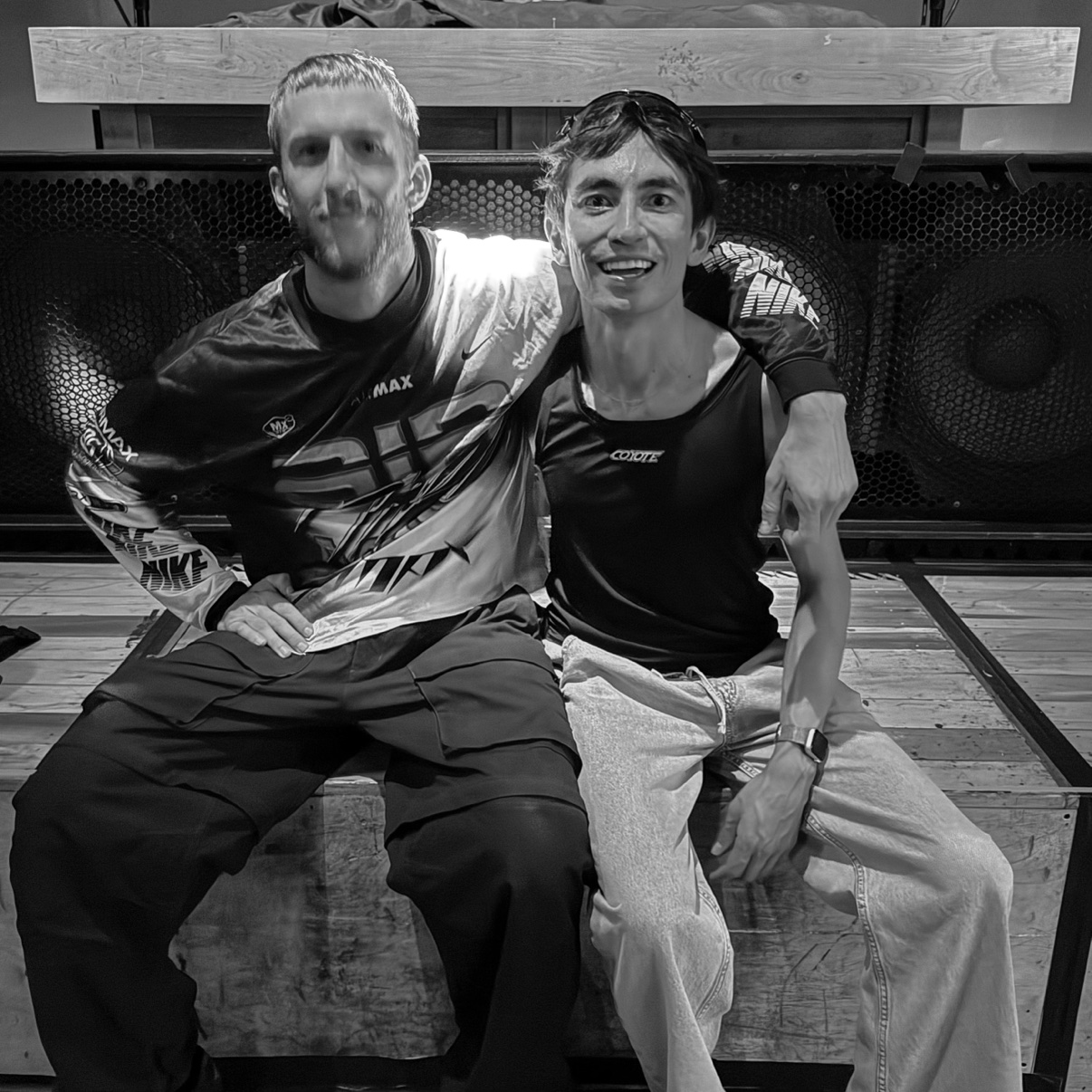 Black-and-white photo of Eldor, right, smiling, seated with a DJ in front of the speaker stacks