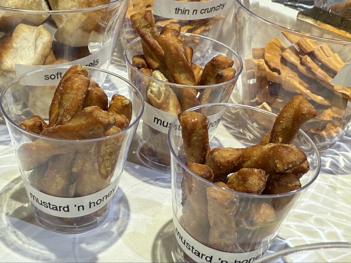 Clear cups of pretzels on a table, labeled mustard 'n honey, soft, and thin n crunchy