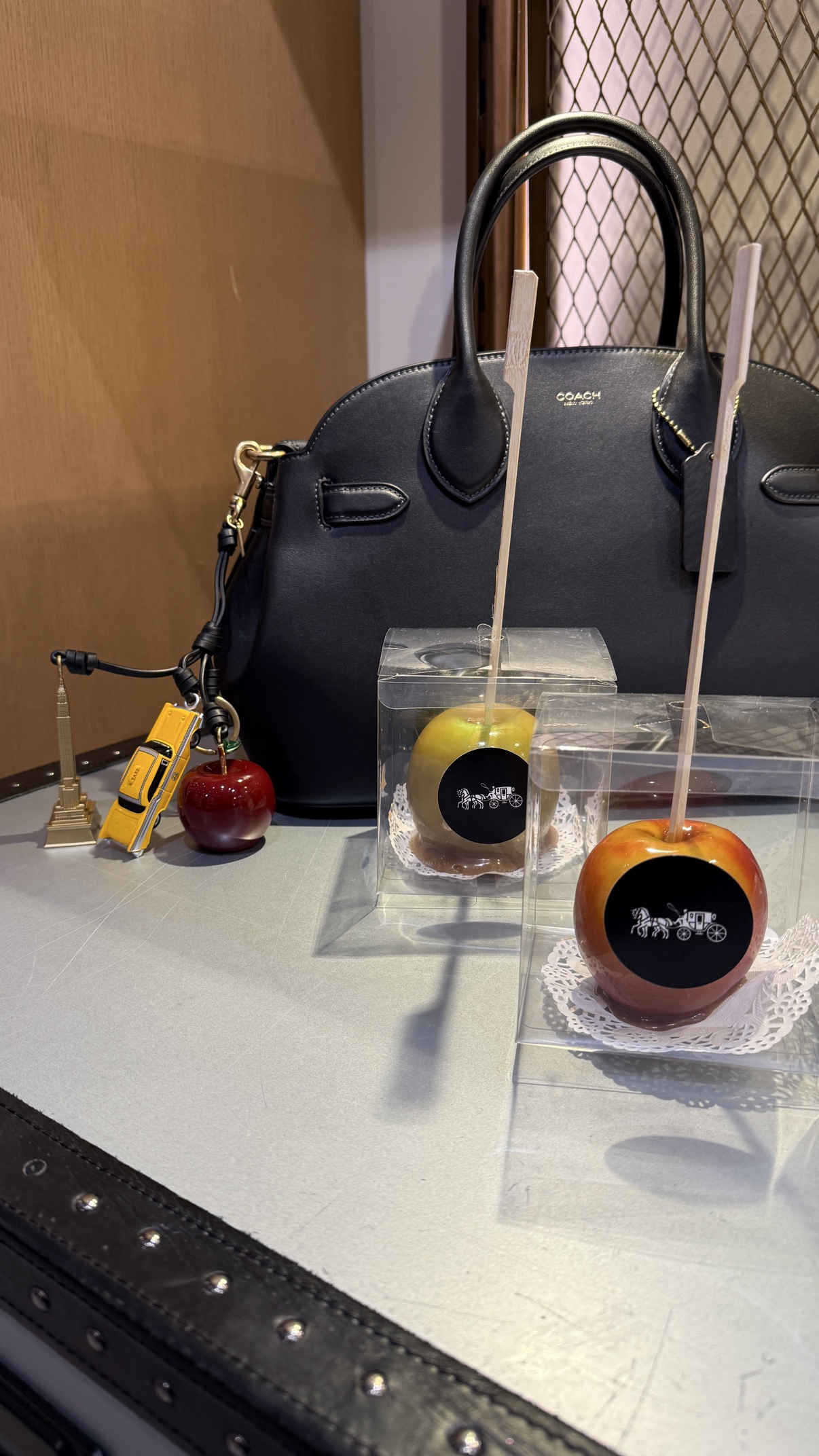 Caramel apples in clear gift boxes beside a black Coach bag with New York charms — a taxi, the Empire State Building, and an apple