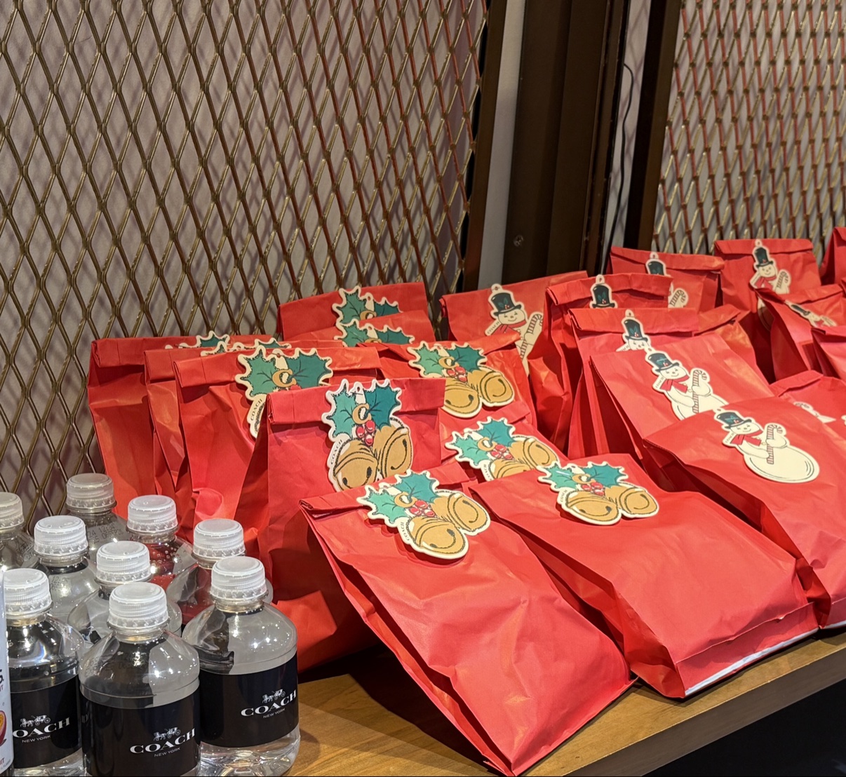 Red holiday gift bags with snowman and holly stickers lined up beside Coach water bottles