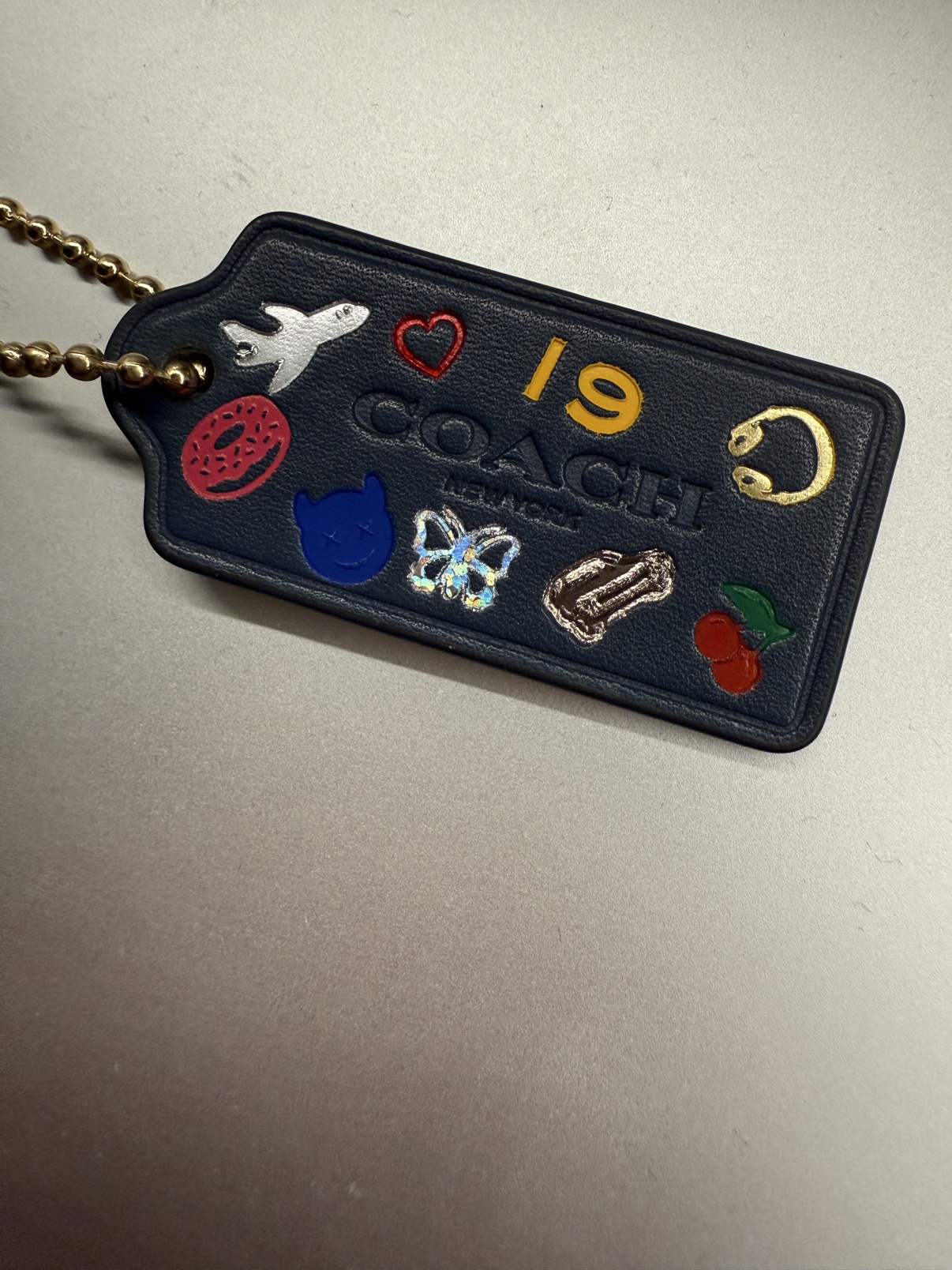 Navy Coach hangtag covered in stamped icons — a plane, a heart, 19, a donut, a cat face, a butterfly, and cherries
