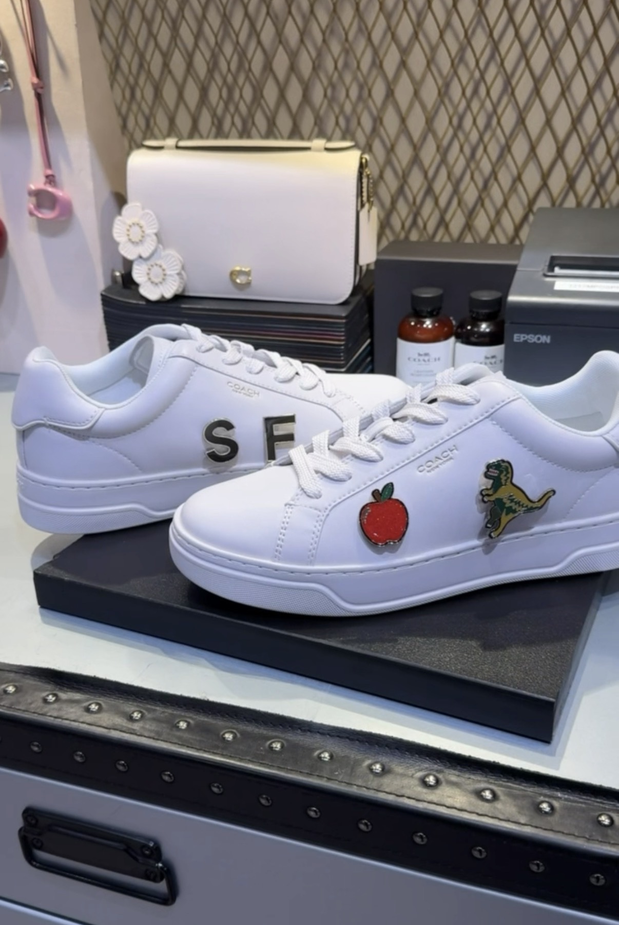 Pair of customized white sneakers with SF letters on one shoe and apple and dinosaur pins on the other