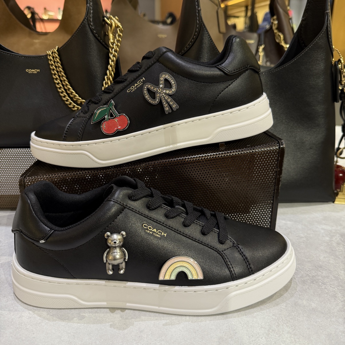 Pair of customized black Coach sneakers — one with cherries and a crystal bow, the other with a metal bear and a rainbow