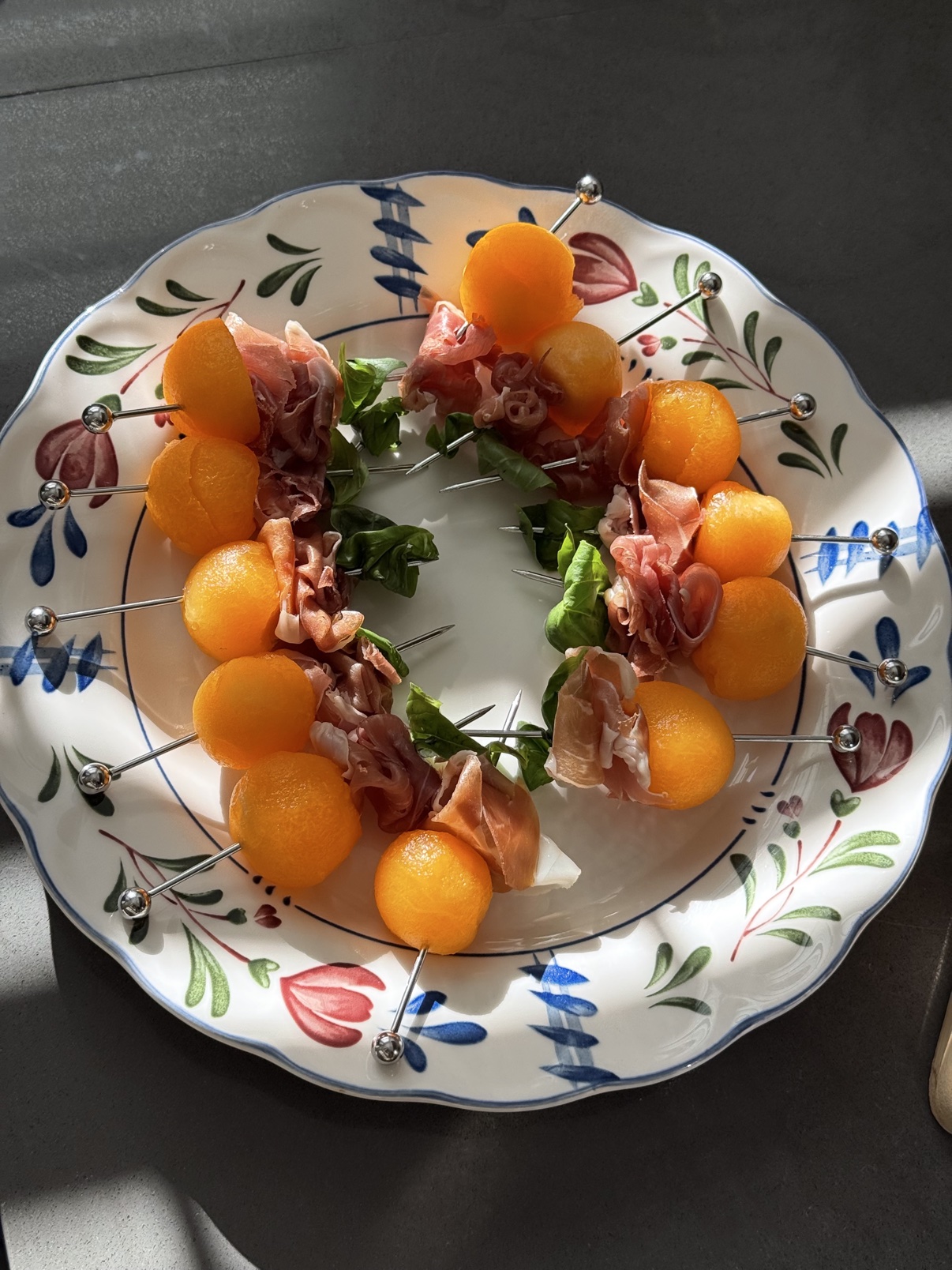 Melon and prosciutto hand-bite skewers with basil, arranged on a floral plate