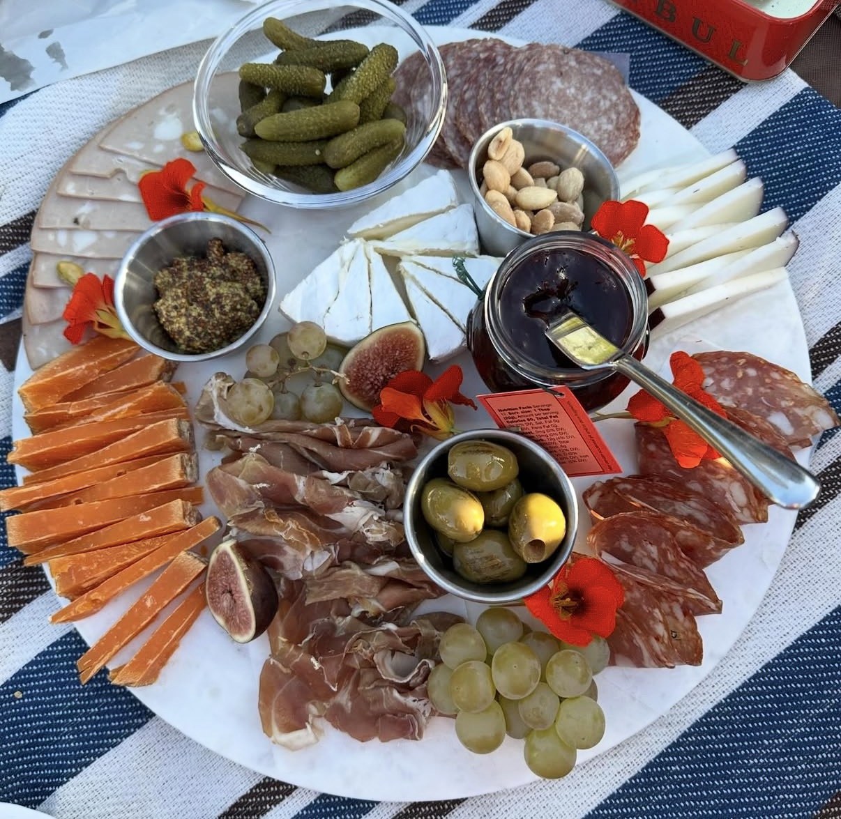 A full charcuterie board on a picnic blanket — cheeses, salami, prosciutto, cornichons, olives, figs, grapes and jam