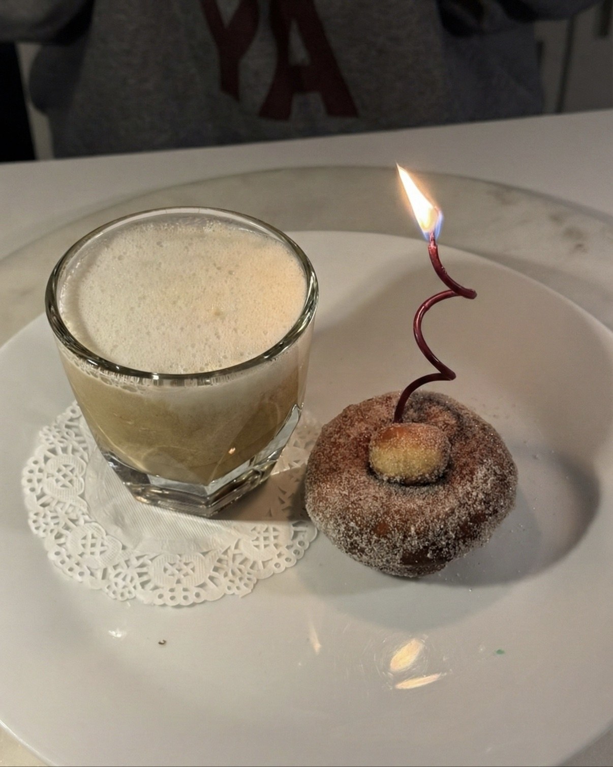 A glass of semifreddo cappuccino beside a cinnamon-sugar donut with a lit candle, on a white plate